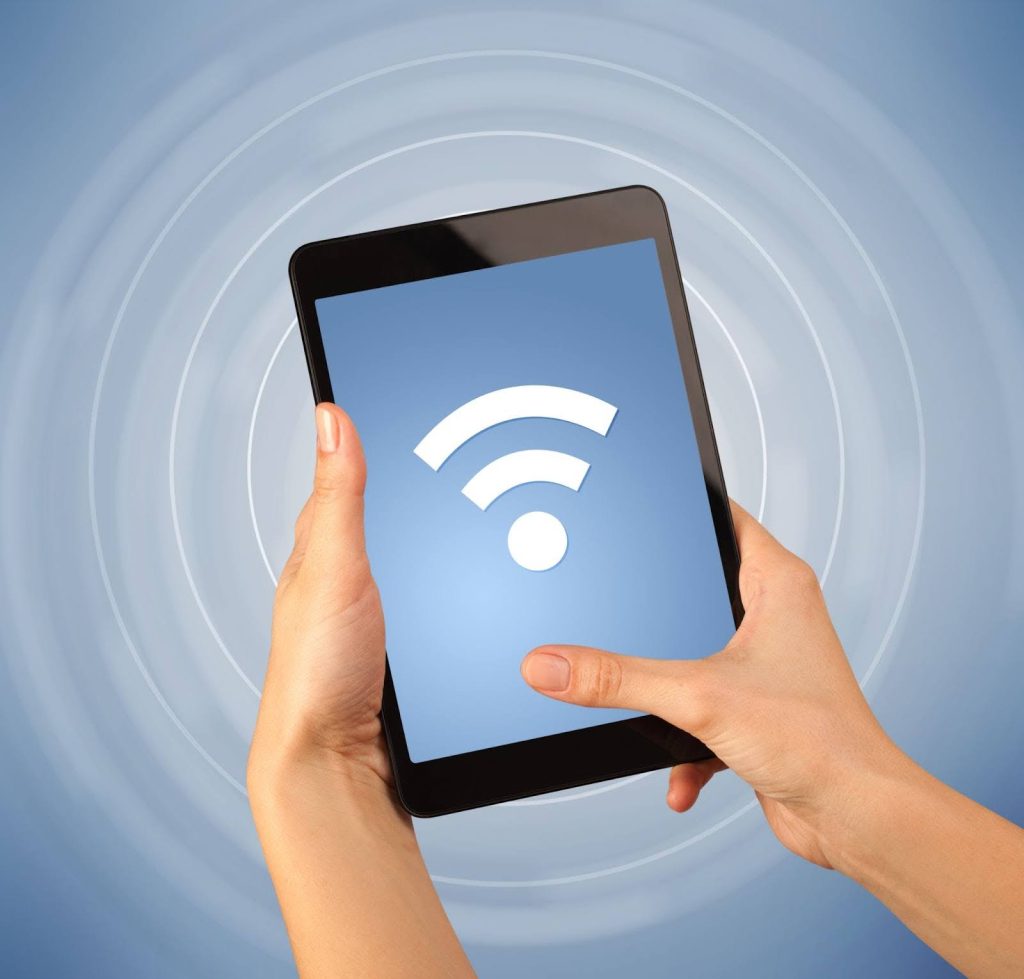 Close-up of hands holding a tablet displaying a large white Wi-Fi symbol on the screen, set against a blue background with radiating signal wave patterns